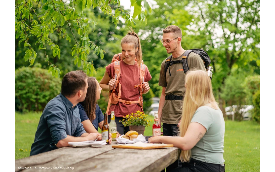 Zwei Wanderer treffen auf drei junge Leute, die gerade an einem Tisch eine Pause machen