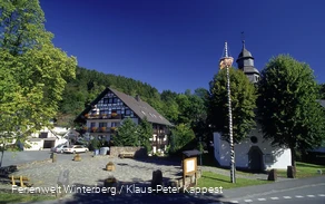 Ein gepflasterter Platz mit Sitzgelegenheiten und Informationstafeln neben einer Kapelle.