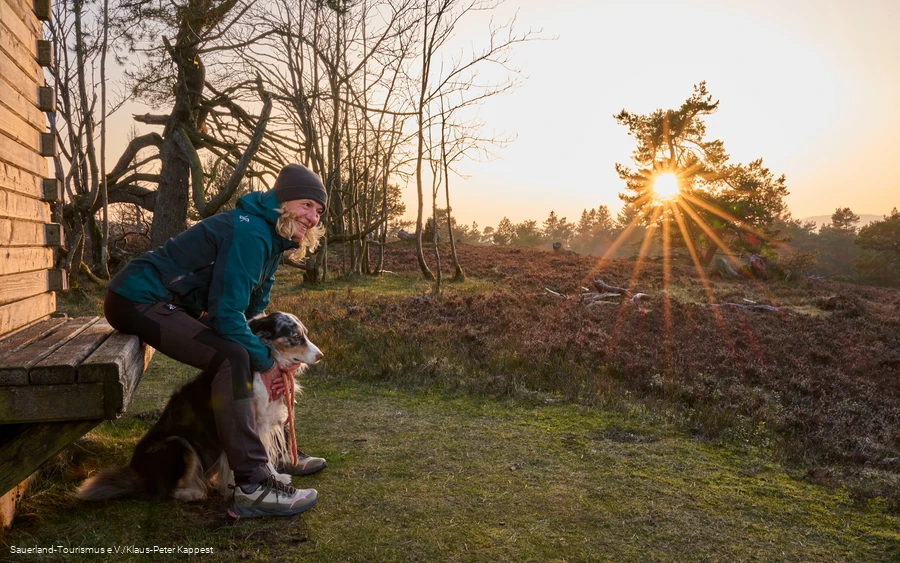 Eine Frau sitzt mit einem Hund an einer Hütte im Sonneuntergang auf der Kahlen Pön in Willingen