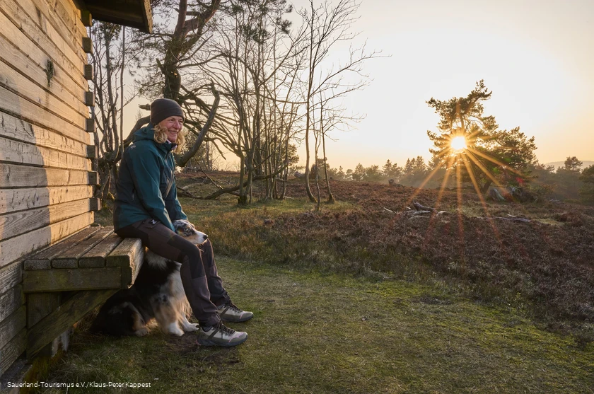 Auf einer Bank an einer H&uuml;tte auf dem Kahlen P&ouml;n sitzt Frau mit Hund im Winterlicht